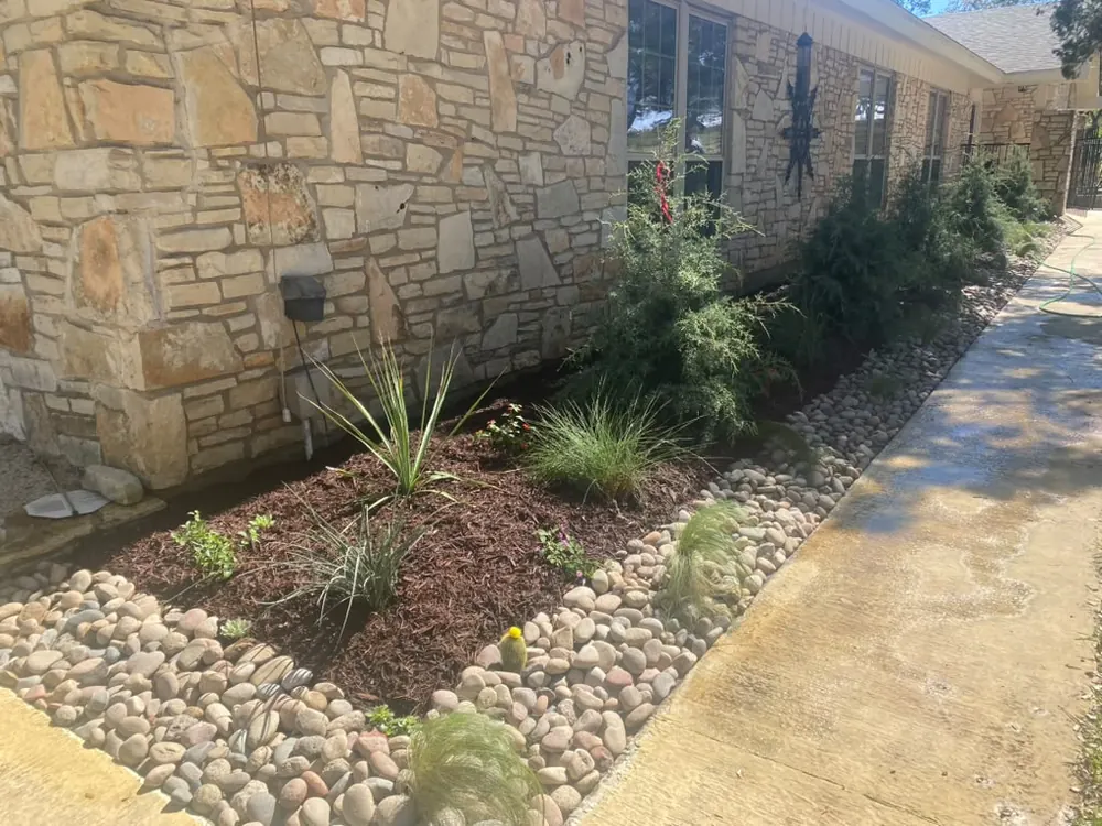 River rock and planting beds alongside a limestone wall and concrete walkway